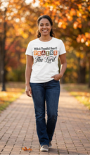 Load image into Gallery viewer, Smiling woman wearing a white faith tee reading With A Thankful Heart I PRAISE The Lord in retro multicolor block letters and script, walking on a fall path