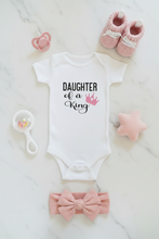 Load image into Gallery viewer, A baby bodysuit laid flat on a white marble surface, printed with the words 'DAUGHTER of a King' in black with a pink crown graphic. Surrounding the bodysuit are pink baby accessories including a pacifier, knit booties, a rattle, a star toy, and a bow headband.
