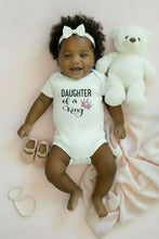 Load image into Gallery viewer, A white short-sleeve baby bodysuit laid flat on a solid red background. The bodysuit features the text 'DAUGHTER of a King' in black, with a small pink crown graphic.
