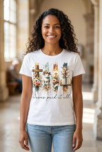 Load image into Gallery viewer, Smiling woman wearing a white Jesus Paid It All graphic tee with a floral cross, lamb, and open Bible design, standing in a bright church foyer
