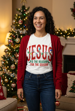 Load image into Gallery viewer, Smiling woman wearing a white Christmas graphic tee with bold red JESUS lettering, colorful Christmas lights, and holly, standing by a decorated Christmas tree
