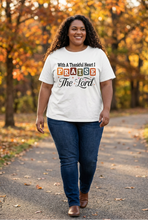 Load image into Gallery viewer, Smiling plus-size woman with long dark curls wearing a white faith tee reading With A Thankful Heart I PRAISE The Lord in retro multicolor block letters and script, walking a fall path
