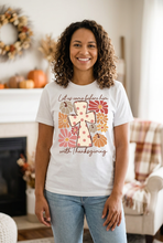 Load image into Gallery viewer, Smiling woman wearing a white Thanksgiving graphic tee with a polka-dot cross, autumn flowers, and pumpkins, in a cozy fall living room
